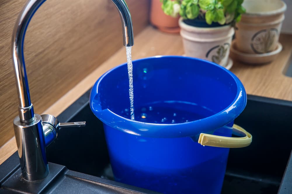 Blauer Eimer in einer Spüle wird mit ablaufendem Trinkwasser befüllt.